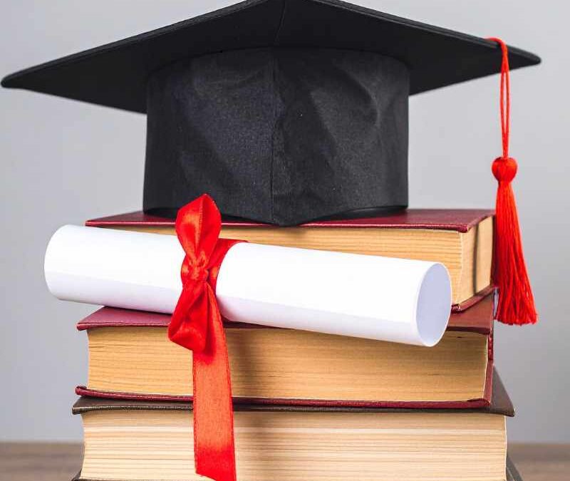 A black graduation cap with a red tassel sits on top of a stack of four books, with a rolled-up white diploma tied with a red ribbon resting against them - www.integratedinsurancesc.com